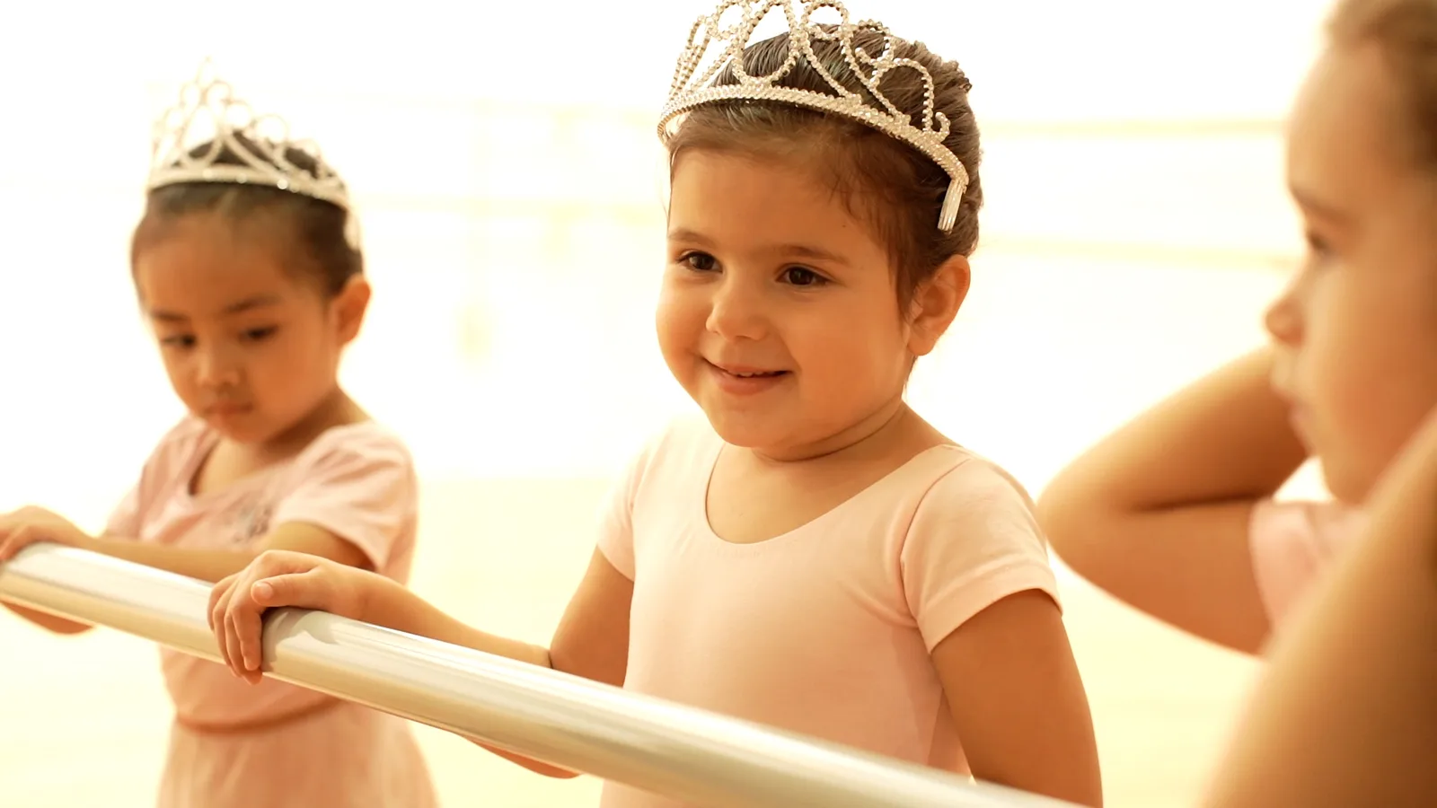 Preschool girl in a tiara and pink leotard smiling at the ballet barre