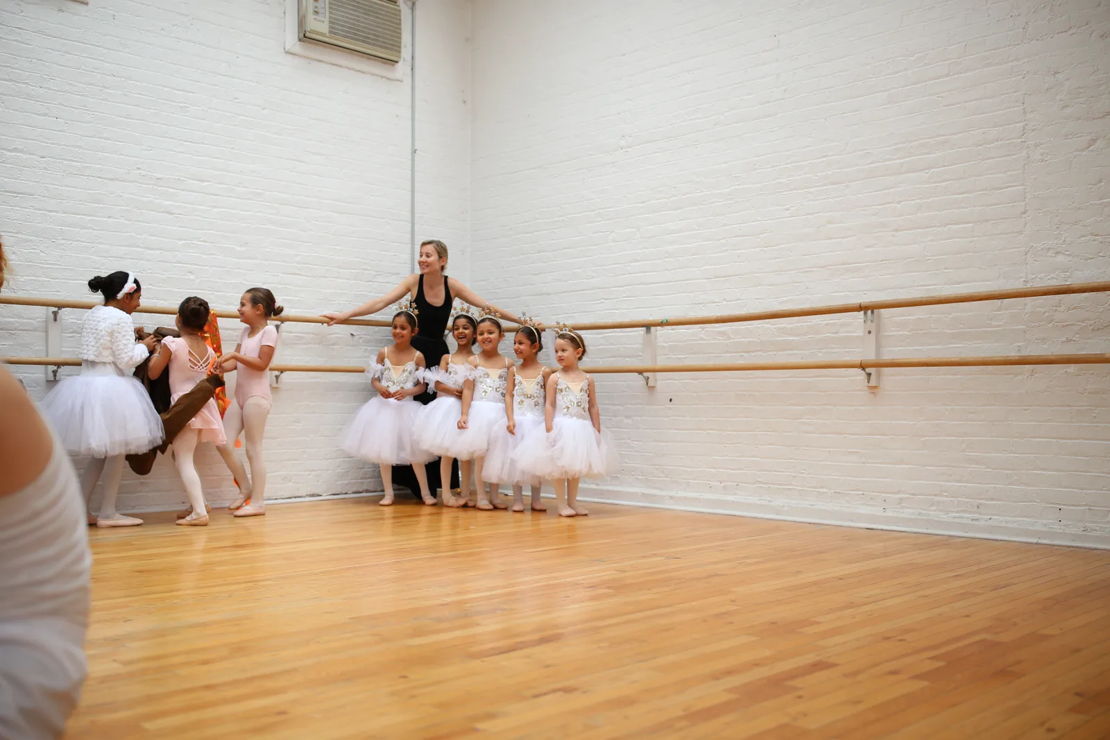 Row of young dancers in white tutus lined up at the barre with their teacher