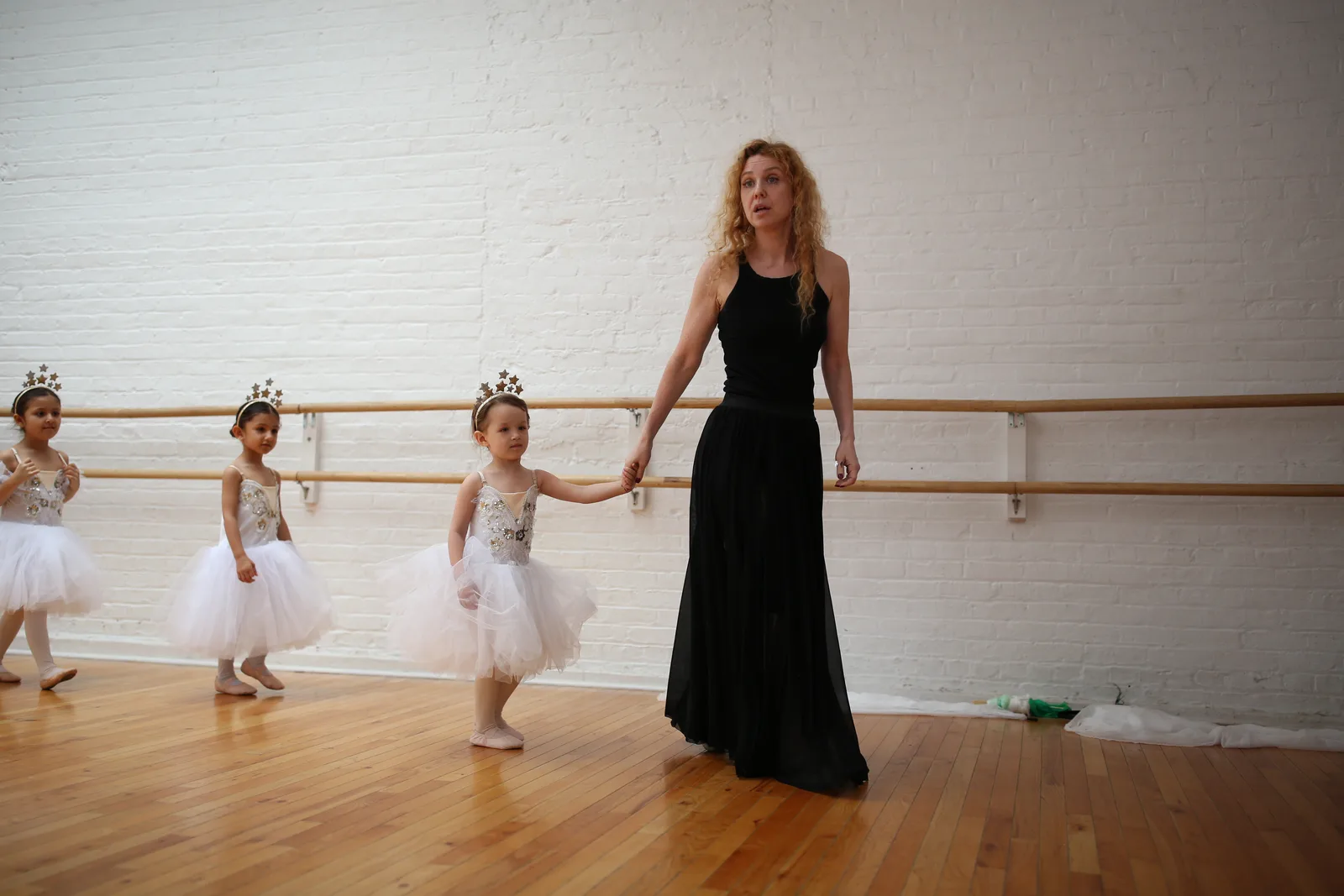 Teacher leading a small child by the hand past the barre while other young dancers wait