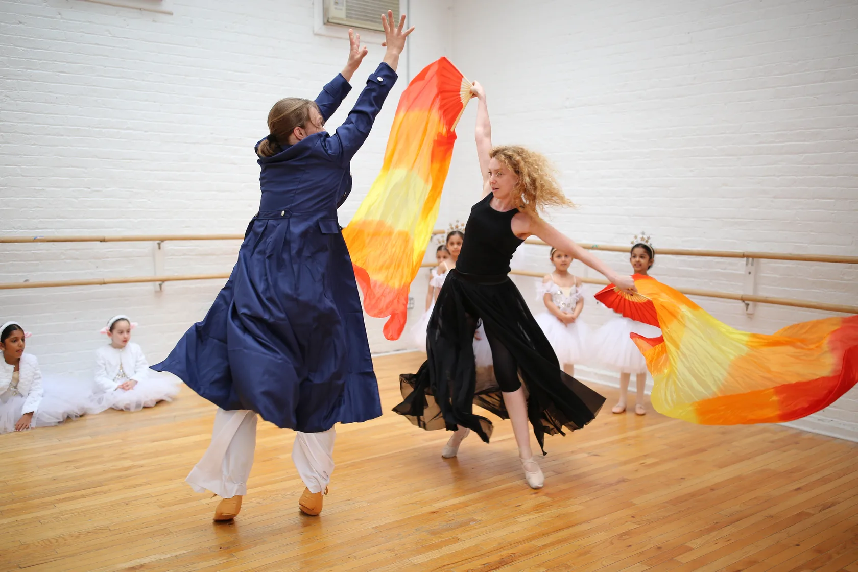 Two teachers rehearsing with fire-coloured silk fans while students watch from the barre