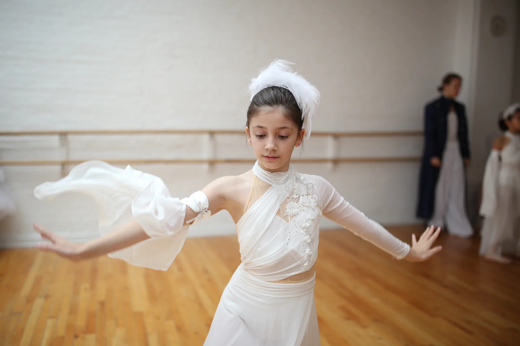 A young dancer in a white costume and feather headpiece opening her arms in rehearsal