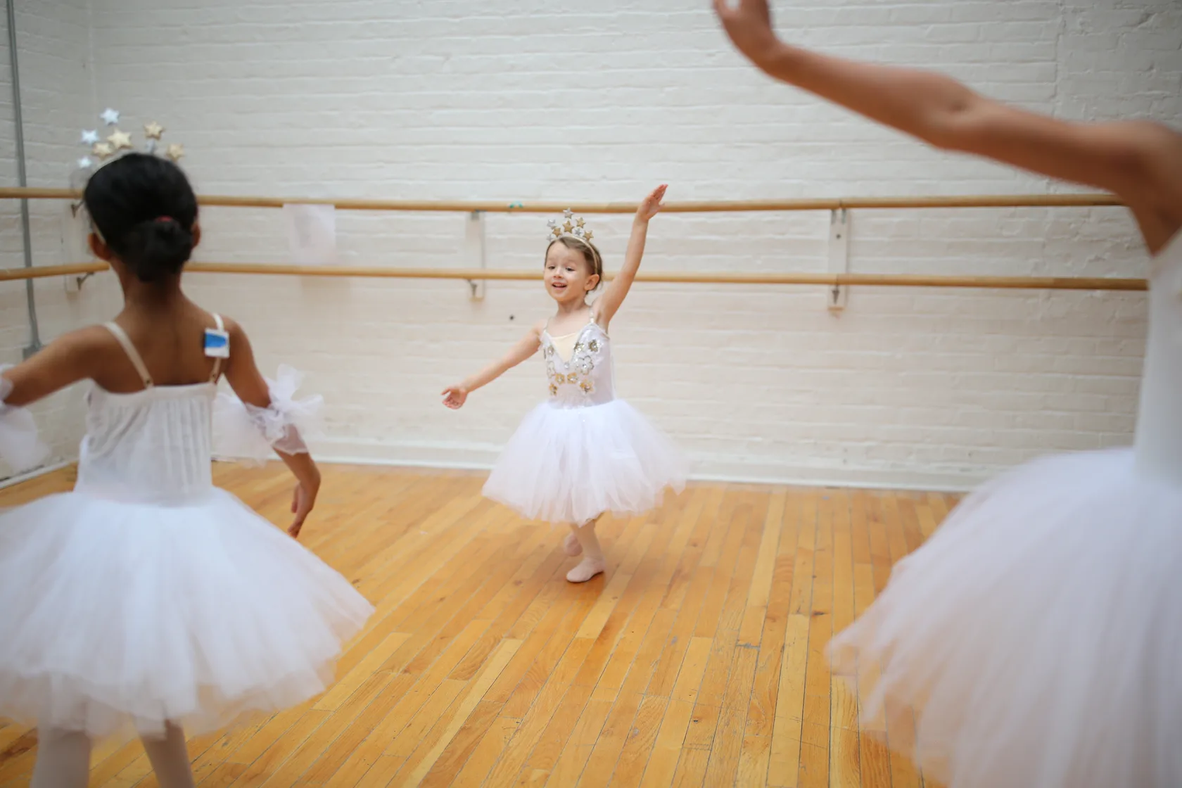 A young dancer in a star crown spinning with one arm raised as classmates dance around her